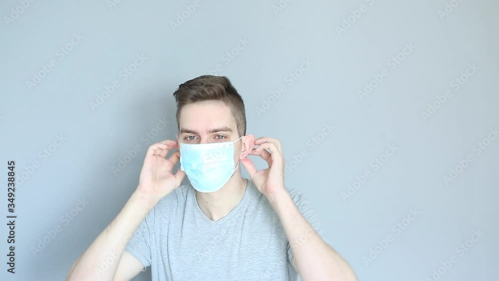 Young sick man in grey shirt on grey background puts on medical mask Young sick man in grey shirt on grey background puts on medical mask