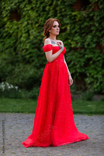 Attractive redhead tattooed woman in red dress and diadema posing on blurred medieval castle background