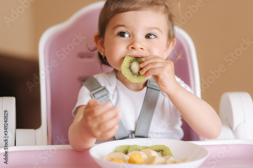 Portrait of happy little kid eating kiwi in high chair. Healthy nutrition for kids