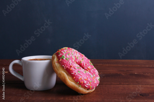 Donut with pink glaze and cup of black coffee on brown wooden table against dark blue wall background. Side view, copy space, close-up. Bakery, coffee shop menu, breakfast, break concept