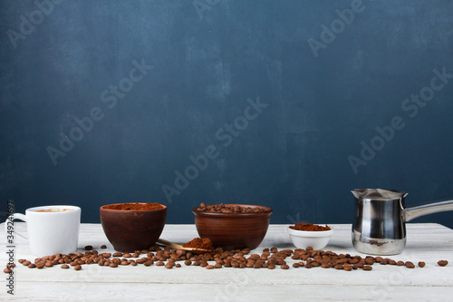 Espresso cup, metal Turkish pot, roasted Arabica beans in clay bowls, coffee powder on white table against dark blue wall. Side view, copy space. Coffee shop, morning, baristas workplace concept