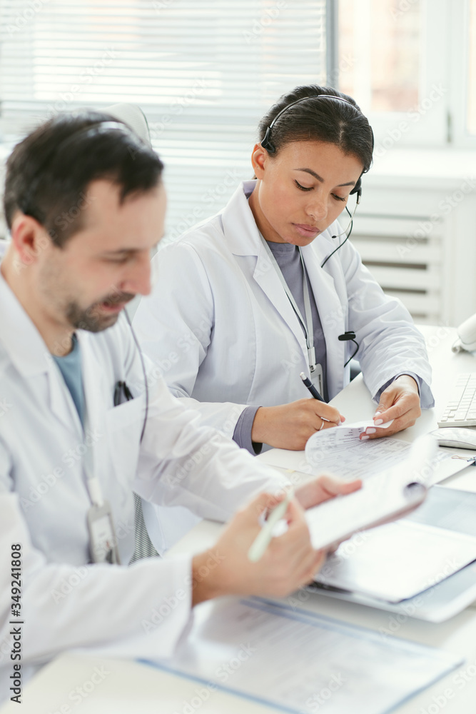 Two serious doctors sitting at the table and doing paperwork together at office Two serious doctors sitting at the table and doing paperwork together at office