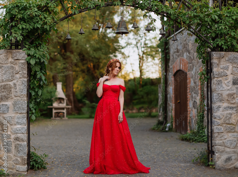 Attractive redhead tattooed woman in red dress and diadema on blurred medieval castle background Attractive redhead tattooed woman in red dress and diadema on blurred medieval castle background