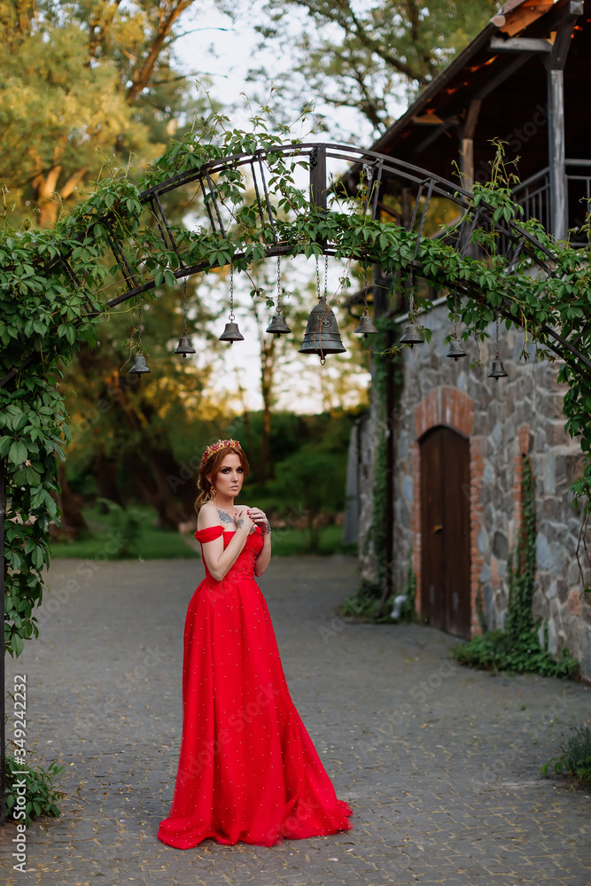 Attractive redhead tattooed woman in red dress and diadema on blurred medieval castle background