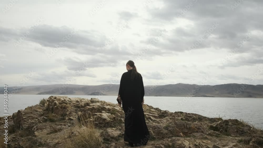 beautiful woman in black dress walks to edge of cliff at top of mountain.view from behind, view of large lake.young woman enjoys mountain scenery and windy weather beautiful woman in black dress walks to edge of cliff at top of mountain.view from behind, view of large lake.young woman enjoys mountain scenery and windy weather
