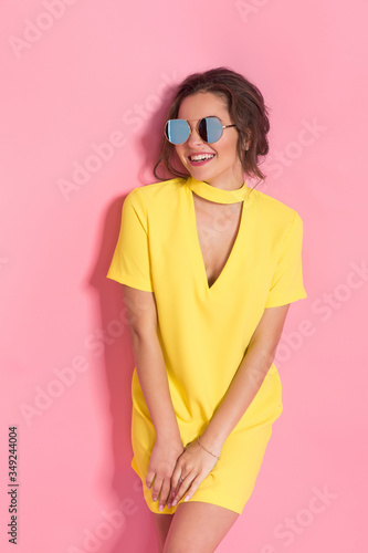Beautiful girl wearing yellow dress and sunglasses posing on pink background in studio. © zamuruev