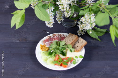 Appetizer plate with ham, cheese, green basil, cilantro, cucumber, tomato, bread, mustard on black wooden background with bird cherry flowers
