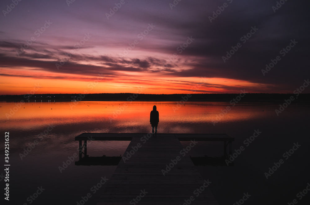 Silhouette of a man standing on a wooden pier in the lake against the background of an incredible colorful bright sunset. Amazing nature.