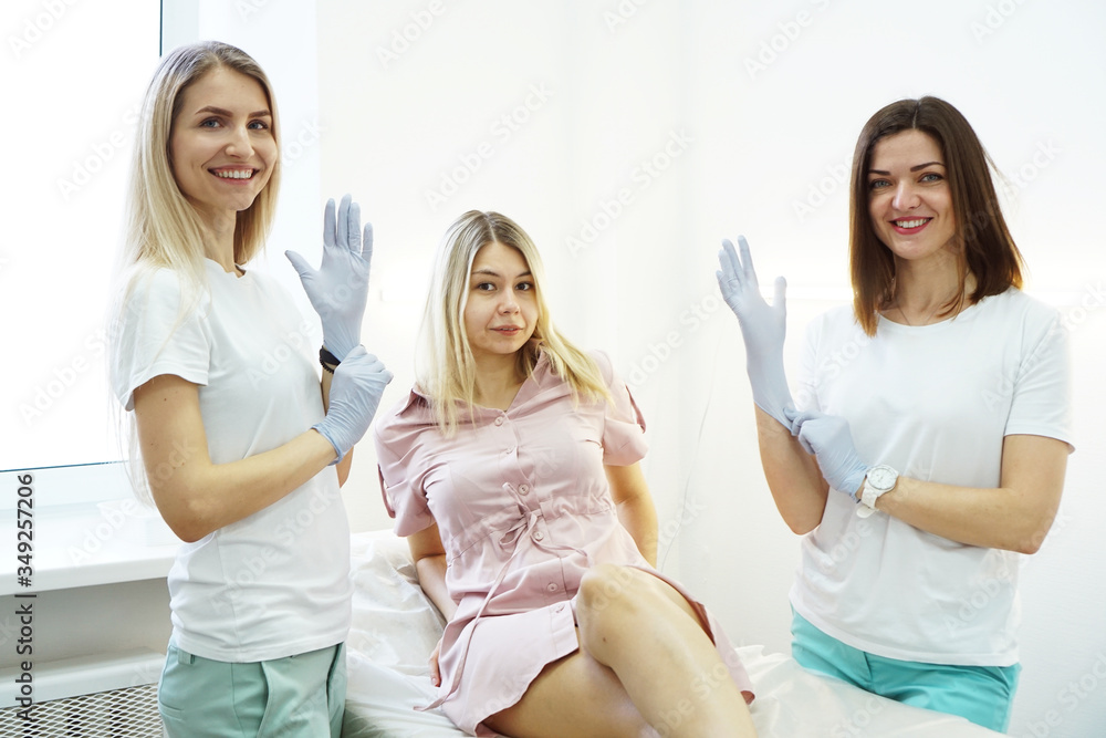 Two young beauticians or doctors put a glove on their hand and smile. The patient lies on the couch and is afraid or surprised