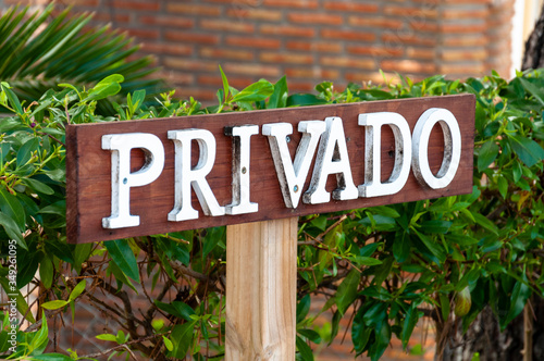 Wooden plate with white letters "PRIVADO" - private on the background of green bushes and a brick wall.