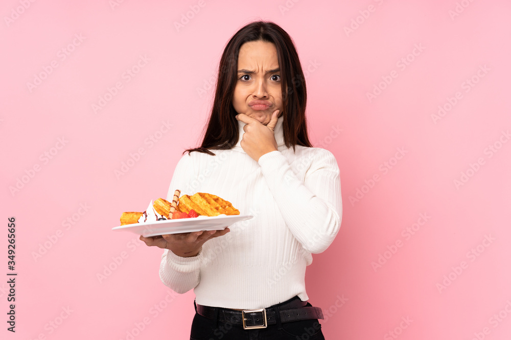Young brunette woman holding waffles over isolated pink background thinking