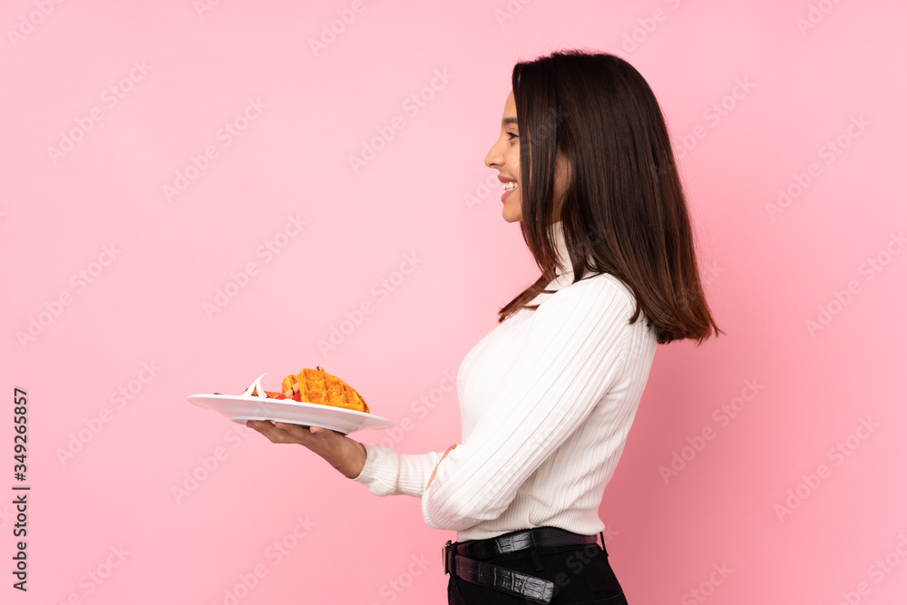 Young brunette woman holding waffles over isolated pink background in lateral position Young brunette woman holding waffles over isolated pink background in lateral position