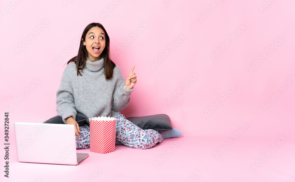 Young mixed race woman eating popcorn while watching a movie on the laptop intending to realizes the solution while lifting a finger up Young mixed race woman eating popcorn while watching a movie on the laptop intending to realizes the solution while lifting a finger up