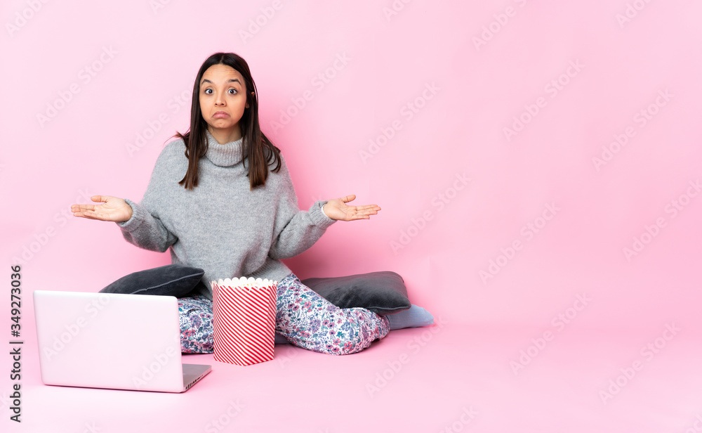 Young mixed race woman eating popcorn while watching a movie on the laptop making doubts gesture Young mixed race woman eating popcorn while watching a movie on the laptop making doubts gesture