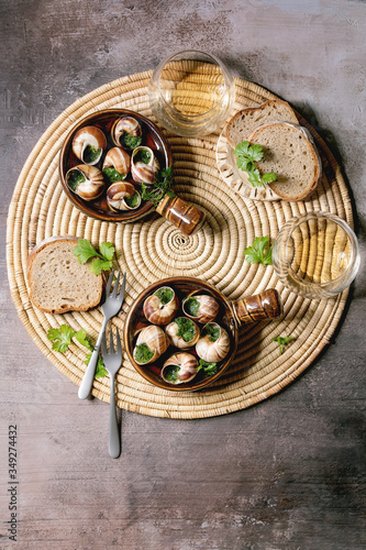 Escargots de Bourgogne - Snails with herbs butter, gourmet dish, in traditional ceramic pans with coriander, bread, glass of white wine on straw napkin over brown textured background. Flat lay, space