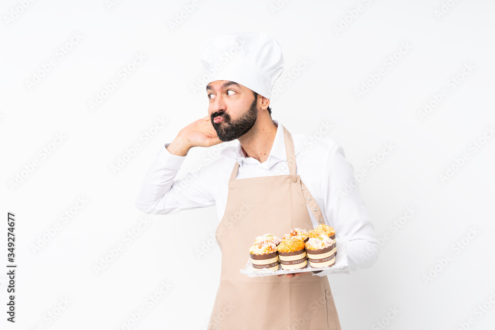 Young man holding muffin cake over isolated white background listening to something by putting hand on the ear Young man holding muffin cake over isolated white background listening to something by putting hand on the ear