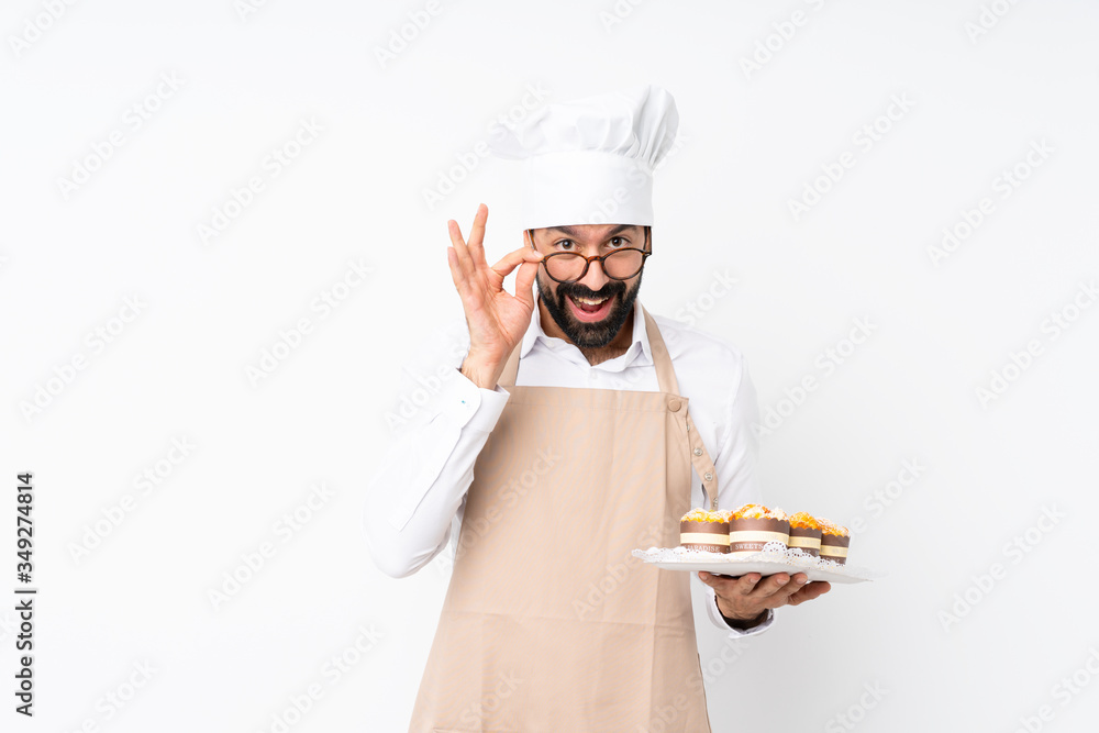 Young man holding muffin cake over isolated white background with glasses and surprised Young man holding muffin cake over isolated white background with glasses and surprised