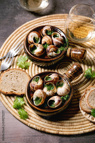 Escargots de Bourgogne - Snails with herbs butter, gourmet dish, in two traditional ceramic pans with coriander, bread and glass of white wine on straw napkin over brown textured background.