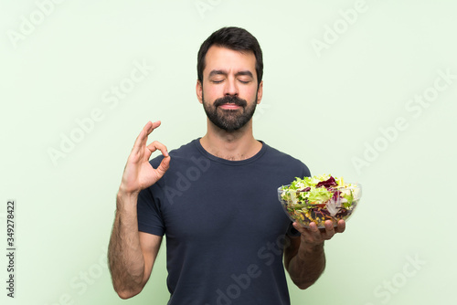 Young handsome man with salad over isolated green wall in zen pose