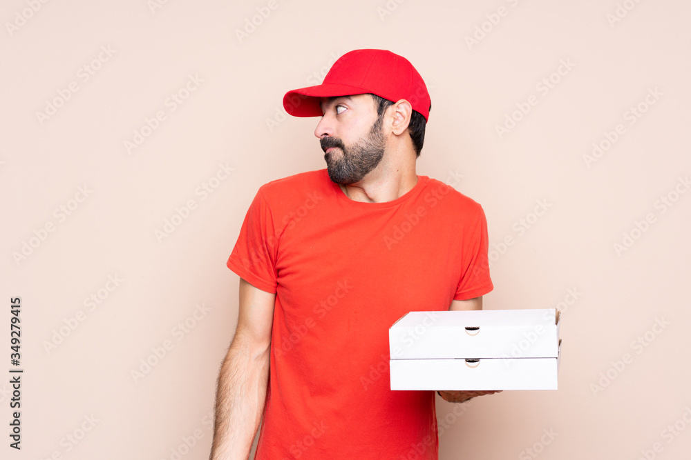 Young man holding a pizza over isolated background making doubts gesture looking side Young man holding a pizza over isolated background making doubts gesture looking side