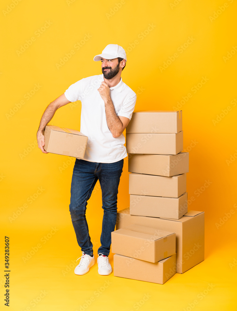 Full-length shot of delivery man among boxes over isolated yellow background scheming something Full-length shot of delivery man among boxes over isolated yellow background scheming something