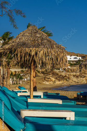 Luxurious beach chairs   umbrellas by an empty sandy beach. Evening view of wooden deck chairs with blue mattresses below bamboo sun protect umbrellas without crowd at Mykonos Greece Paradise Beach.