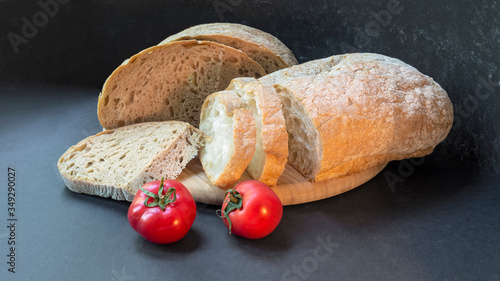 Beautiful bread close-up, two types of bread made from wheat and rye flour and two fresh tomatoes on a round wooden board on a black background. Sliced and half a loaf.
