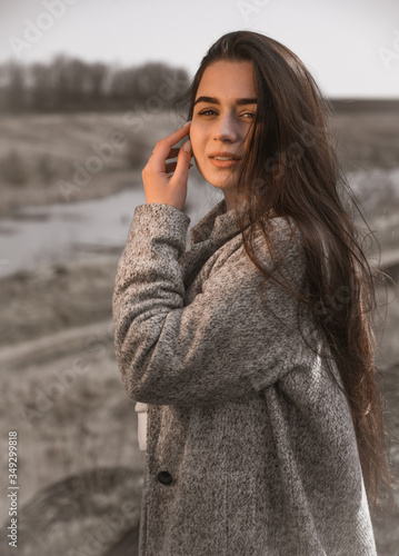 Natural light portrait  neutral real skin tones. Golden hour sun rays brighten face of beautiful young Ukrainian woman wearing grey coat. Hand near head  posing photo session outside.