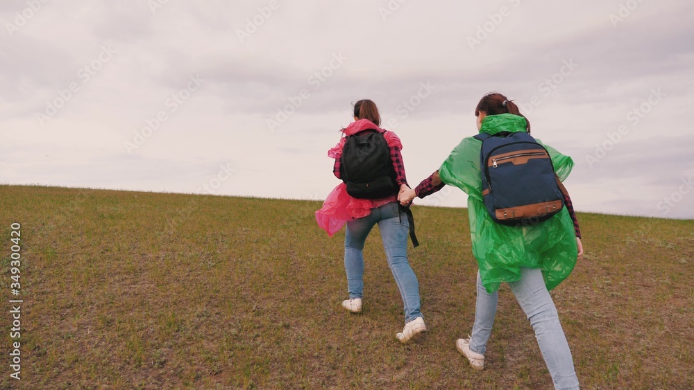 healthy girls tourists travel with backpacks in multi-colored raincoats climbing hill. free women travelers go top of mountain holding hands. slow motion. teamwork travelers. adventure concept.