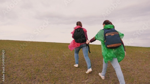 healthy girls tourists travel with backpacks in multi-colored raincoats climbing hill. free women travelers go top of mountain holding hands. slow motion. teamwork travelers. adventure concept.