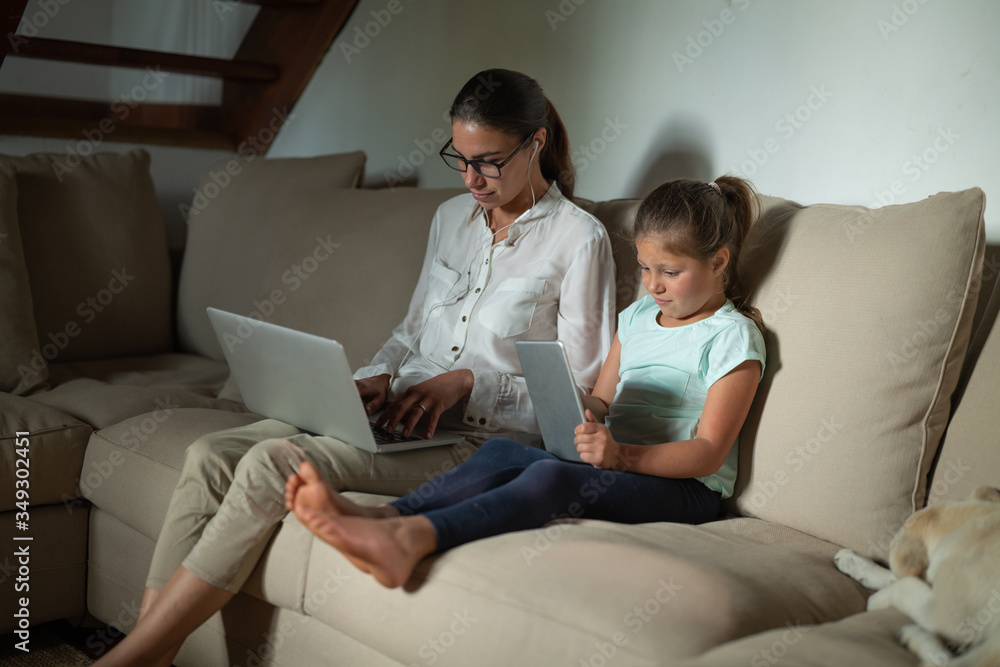 Authentic shot of an young mother is doing smart working with laptop and her daughter is playing with tablet while sitting on a sofa in a living room at home. Authentic shot of an young mother is doing smart working with laptop and her daughter is playing with tablet while sitting on a sofa in a living room at home.