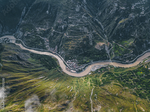 Top down aerial drone view of river and small settlement in between the tall mountains in Tibetan area of Sichuan province, China. Cinematic shot, curved serpentine roads and hills. Journey in spring.