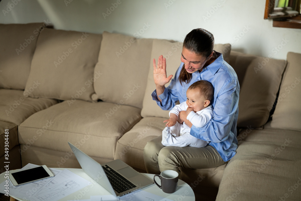 Authentic shot of neo mother and her newborn baby making a video call to father or relatives with laptop on a sofa in a living room. 