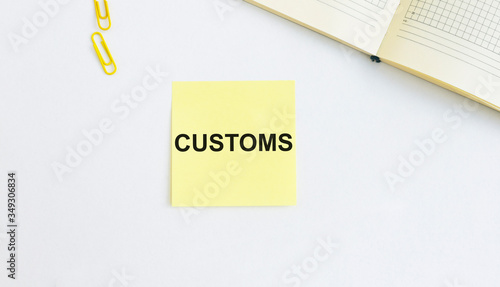 CUSTOMS Word Written on yellow memo note on white table with yellow clips and corner of notebook. Business and government concept