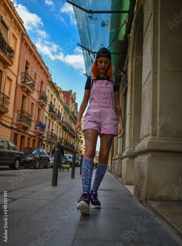 Young woman dressed in a pink denim jumpsuit, cap and very colorful high socks, walking through town alone © Andrea