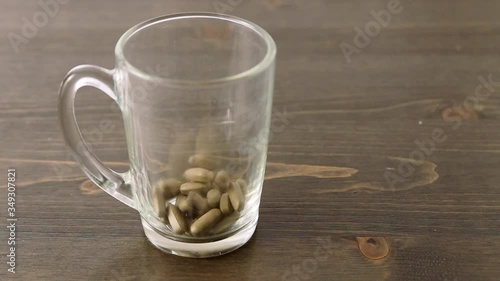 Herbal medicine tablets (black walnut leaves) fall into a transparent empty glass on a wooden table. Concept for self-medication, natural medicine and dangers of overdose
