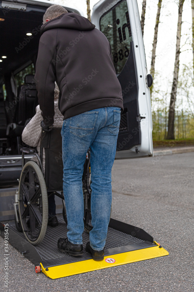 Obraz premium Handicapped person loading in a wheelchair into a minibus for transportation. Male driver and elderly woman in medical masks.