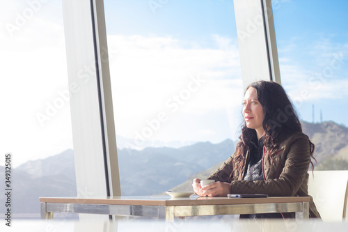 Young woman in cafe looking trough with big windows with blue sky and mountains outdoors