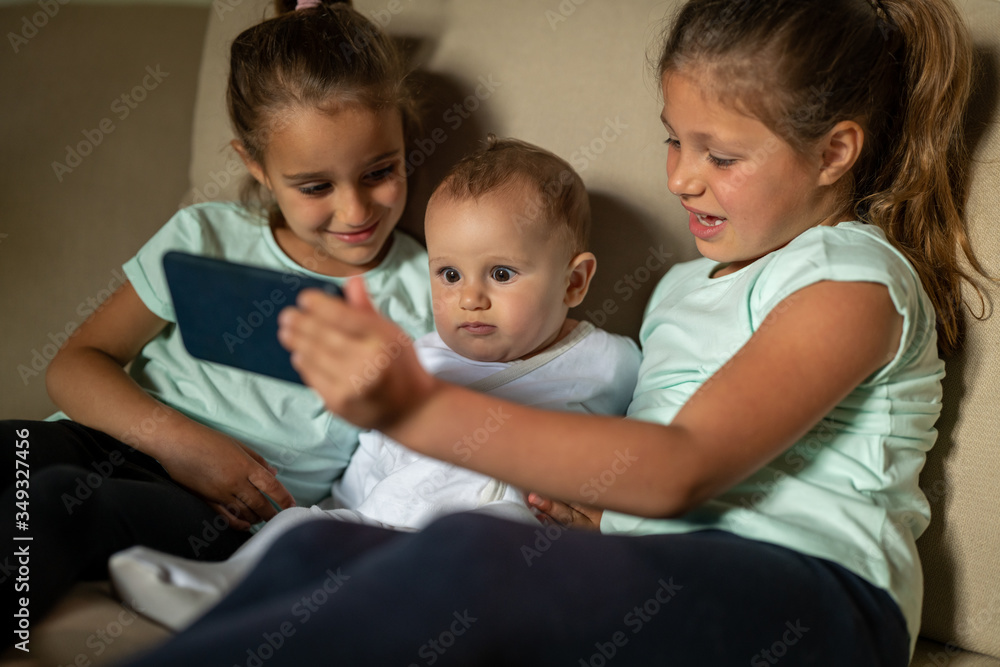 Authentic shot of happy little sisters with newborn brother are making a selfie or video call to parent or relatives while sitting on a sofa in a living room at home. Authentic shot of happy little sisters with newborn brother are making a selfie or video call to parent or relatives while sitting on a sofa in a living room at home.