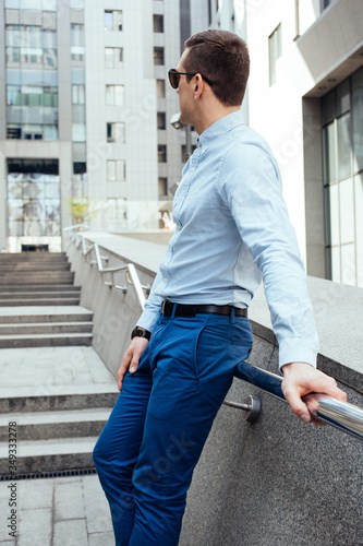 A young man in a blue shirt and sunglasses is standing on the street near the business centre. Waiting for a wife. Business portrait. Stairs on the background. 