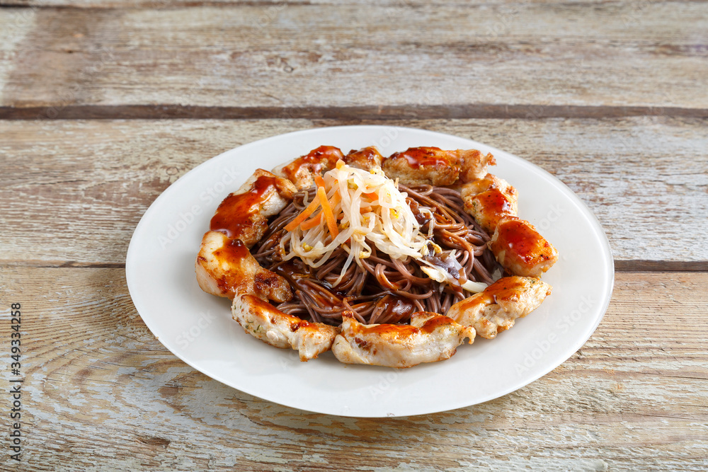 White plate with soba buckwheat noodles and vegetables with chicken on a wooden table. White plate with soba buckwheat noodles and vegetables with chicken on a wooden table.