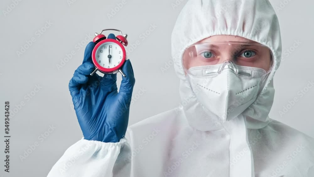 Seasonal disease. Coronavirus pandemic. Female doctor in white ppe googles showing alarm clock. Seasonal disease. Coronavirus pandemic. Female doctor in white ppe googles showing alarm clock.