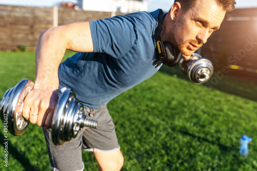 Get in shape without gym. A young guy in sportswear is training in the backyard, he is doing arm and shoulder exercise with iron dumbbells. Concept of healthlife, sport, selfdissipline photo