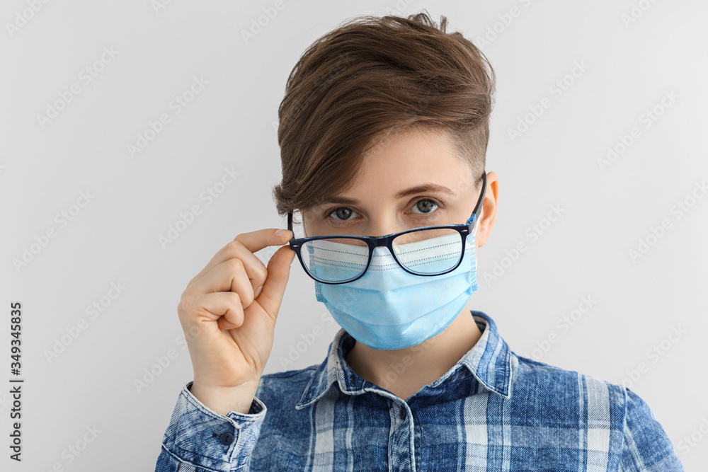 Girl in glasses, a medical mask and a blue plaid shirt. Short hairstyle, European, caucasian woman on a gray background.