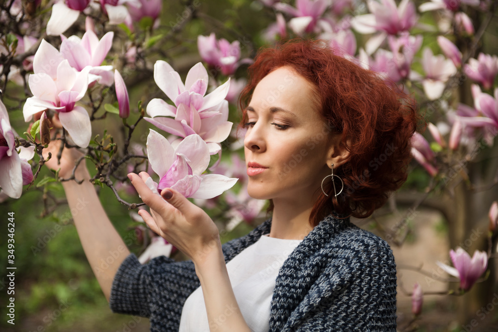 Fototapeta premium Portrait of young pretty red haired woman near blooming magnolia tree on sunny spring day