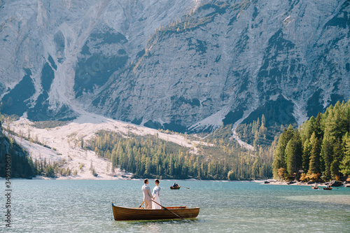 The bride and groom are sailing in a wooden boat with oars at the Lago di Braies in Italy. Wedding in Europe, on Braies lake. Newlyweds are standing embracing in a boat.