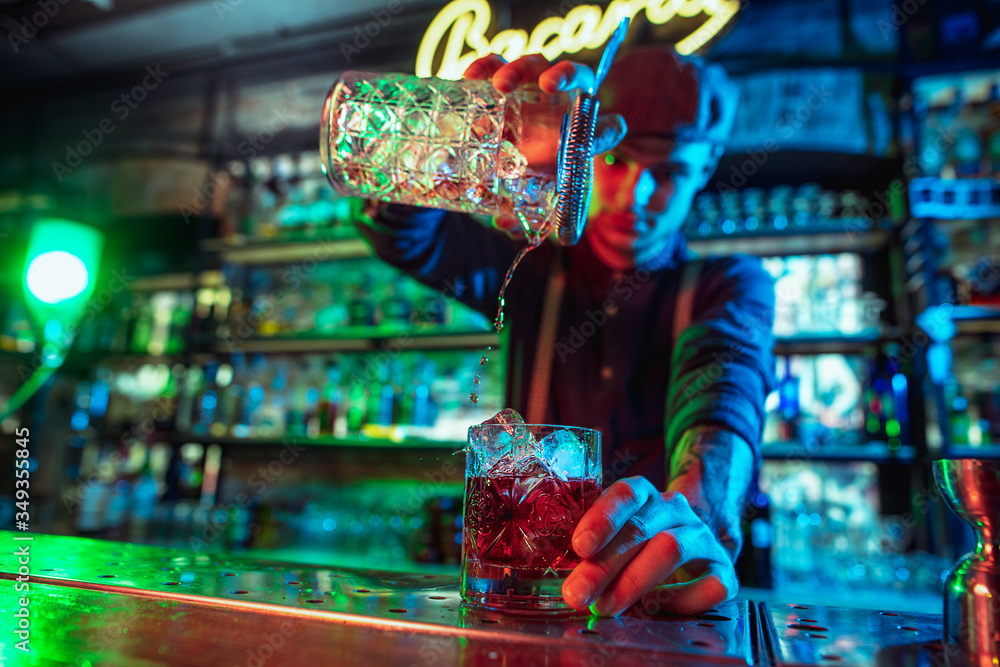 Close up of professional barman finishes preparation of alcoholic cocktail in multicolored neon light, gives it to client. Entertainment, drinks, service concept. Modern bar, trendy neoned colors.