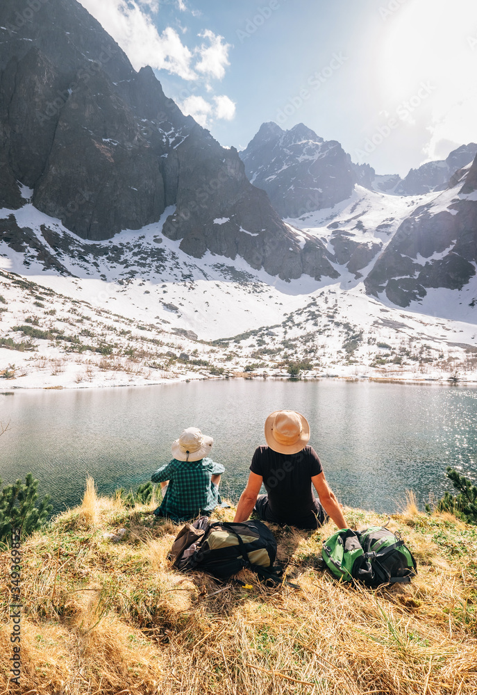 Father and son backpackers resting near the mountain lake Zelene Pleso in Slovakia and enjoying snowy peaks. Spring-summer hiking with kids concept image.