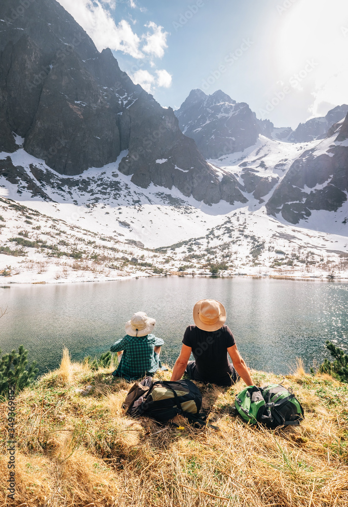 Father and son backpackers resting near the mountain lake Zelene Pleso in Slovakia and enjoying snowy peaks. Spring-summer hiking with kids concept image. Father and son backpackers resting near the mountain lake Zelene Pleso in Slovakia and enjoying snowy peaks. Spring-summer hiking with kids concept image.