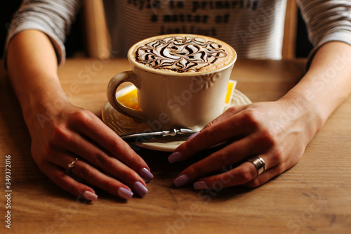 The girl is drinking. The cup of coffee on a background of hands. Girl in a striped sweater with a cup of coffee. A cup of coffee with foam. Delicious patterns on the foam of coffee